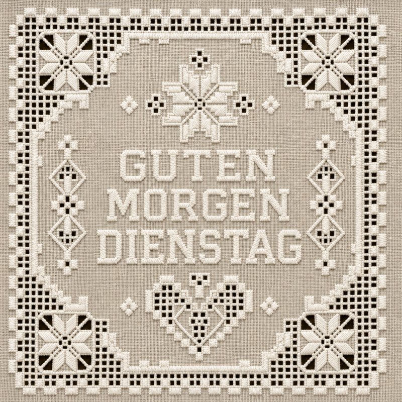 Ein traditioneller Guten-Morgen-Gruß im Kreuzstich-Stil mit Blumenmotiven und dekorativen Mustern auf beigem Stoff.